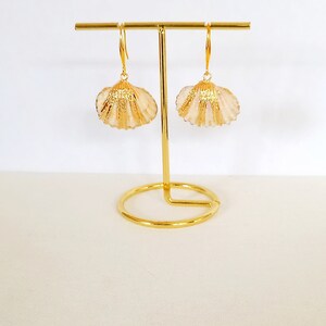 May include: A pair of gold-tone earrings with a seashell design. The earrings are hanging from a gold stand.