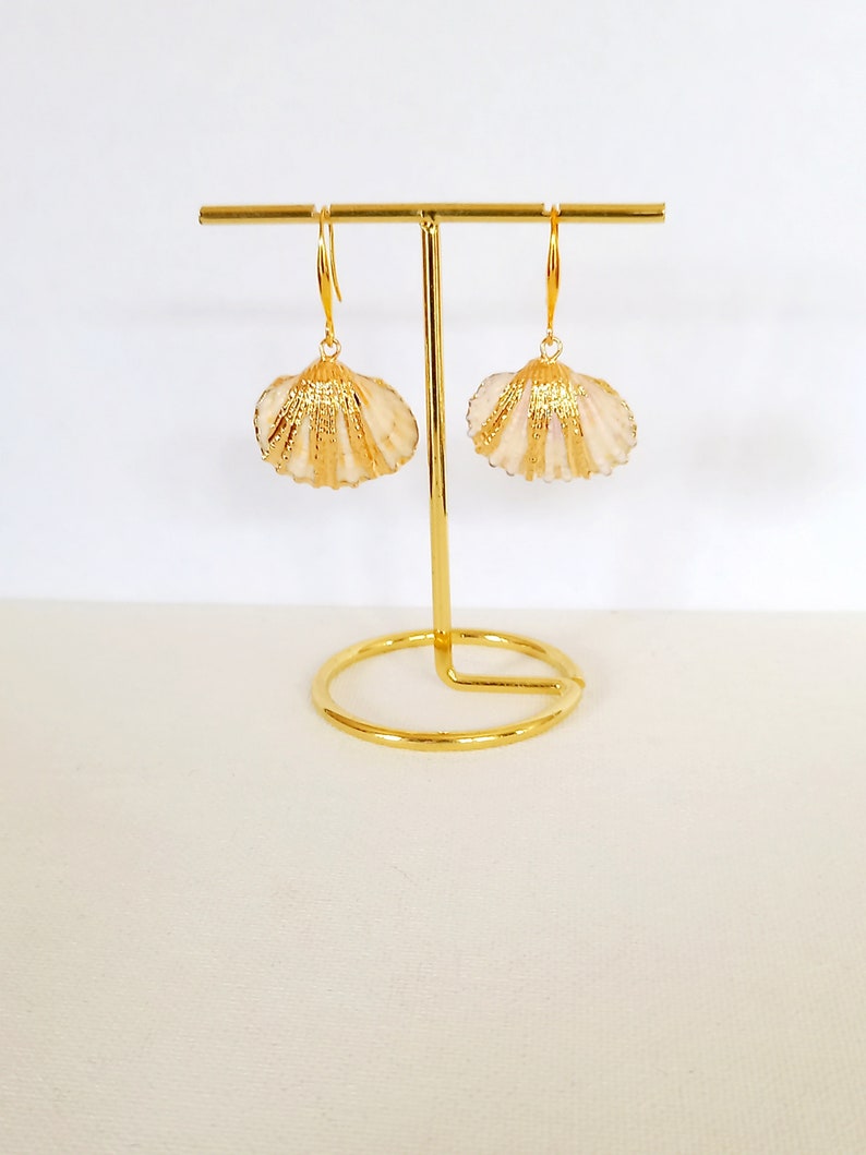 May include: A pair of gold-tone earrings with a seashell design. The earrings are hanging from a gold stand.