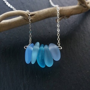 Op de afbeelding: Een zilveren ketting met zes blauwe zeeglas hangers in verschillende tinten blauw. De druppelvormige hangers zijn in een rij gerangschikt. De ketting wordt getoond tegen een donkere achtergrond.
