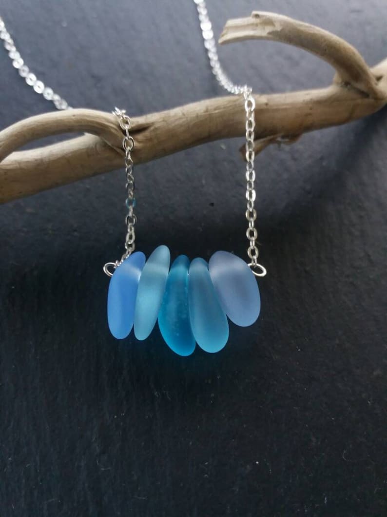 Op de afbeelding: Een zilveren ketting met zes blauwe zeeglas hangers in verschillende tinten blauw. De druppelvormige hangers zijn in een rij gerangschikt. De ketting wordt getoond tegen een donkere achtergrond.