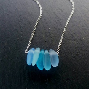 Op de afbeelding: Een zilveren ketting met zes blauwe zeeglas hangers. De hangers hebben verschillende tinten blauw. De ketting is delicaat. De ketting wordt gepresenteerd tegen een donkere achtergrond.