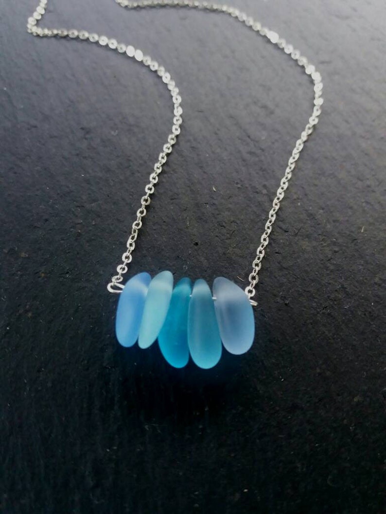 Op de afbeelding: Een zilveren ketting met zes blauwe zeeglas hangers. De hangers hebben verschillende tinten blauw. De ketting is delicaat. De ketting wordt gepresenteerd tegen een donkere achtergrond.