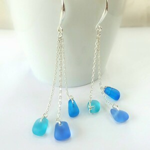 May include: Silver dangle earrings with multiple chains and blue sea glass charms. The sea glass is a gradient of blue colors.