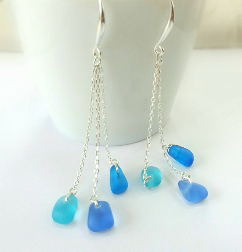 May include: Silver dangle earrings with multiple chains and blue sea glass charms. The sea glass is a gradient of blue colors.