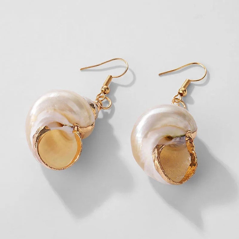 May include: A pair of gold tone earrings featuring a white seashell design. The seashells are detailed with a gold tone trim.