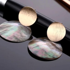 Könnte beinhalten: Ein Paar Ohrringe mit einem goldenen und schillernden Muscheldesign. Die Ohrringe bestehen aus einer runden, goldfarbenen Metallscheibe und einer runden, schillernden Muschelscheibe.
