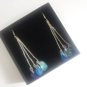 May include: A pair of silver dangle earrings with three blue sea glass stones on each earring. The stones are graduated in size, with the largest at the bottom and the smallest at the top. The earrings are hanging from a black velvet box.