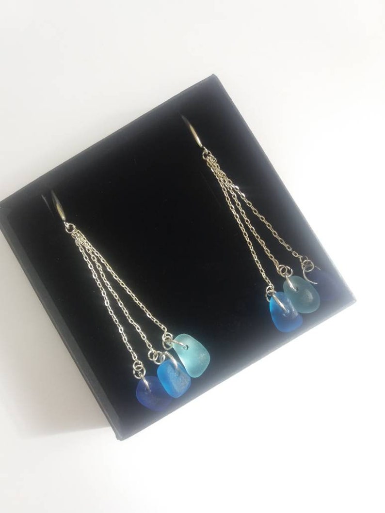 May include: A pair of silver dangle earrings with three blue sea glass stones on each earring. The stones are graduated in size, with the largest at the bottom and the smallest at the top. The earrings are hanging from a black velvet box.