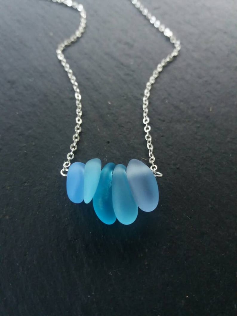 Op de afbeelding: Een zilveren ketting met vijf blauwe zeeglas stukken. Het zeeglas, in blauwtinten, is druppelvormig en horizontaal gerangschikt. De ketting is geplaatst tegen een donkere achtergrond.