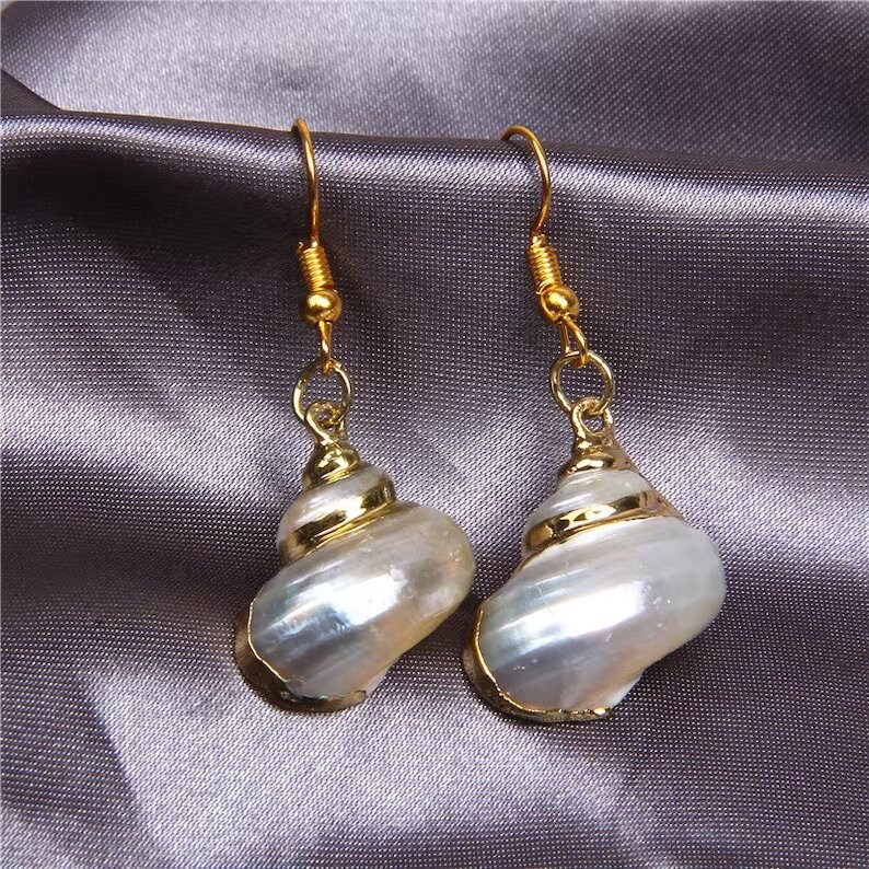 May include: A pair of gold tone earrings featuring a white seashell design. The seashells have a pearlescent finish and are accented with a gold band.
