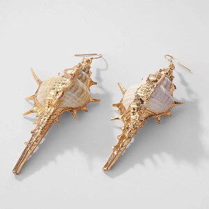 May include: A pair of gold-toned earrings featuring a large, detailed seashell design. The shells are white with brown and gold accents.