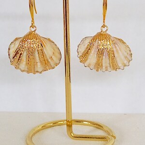 May include: A pair of gold-tone earrings featuring a seashell design. The earrings are hanging from a gold stand.