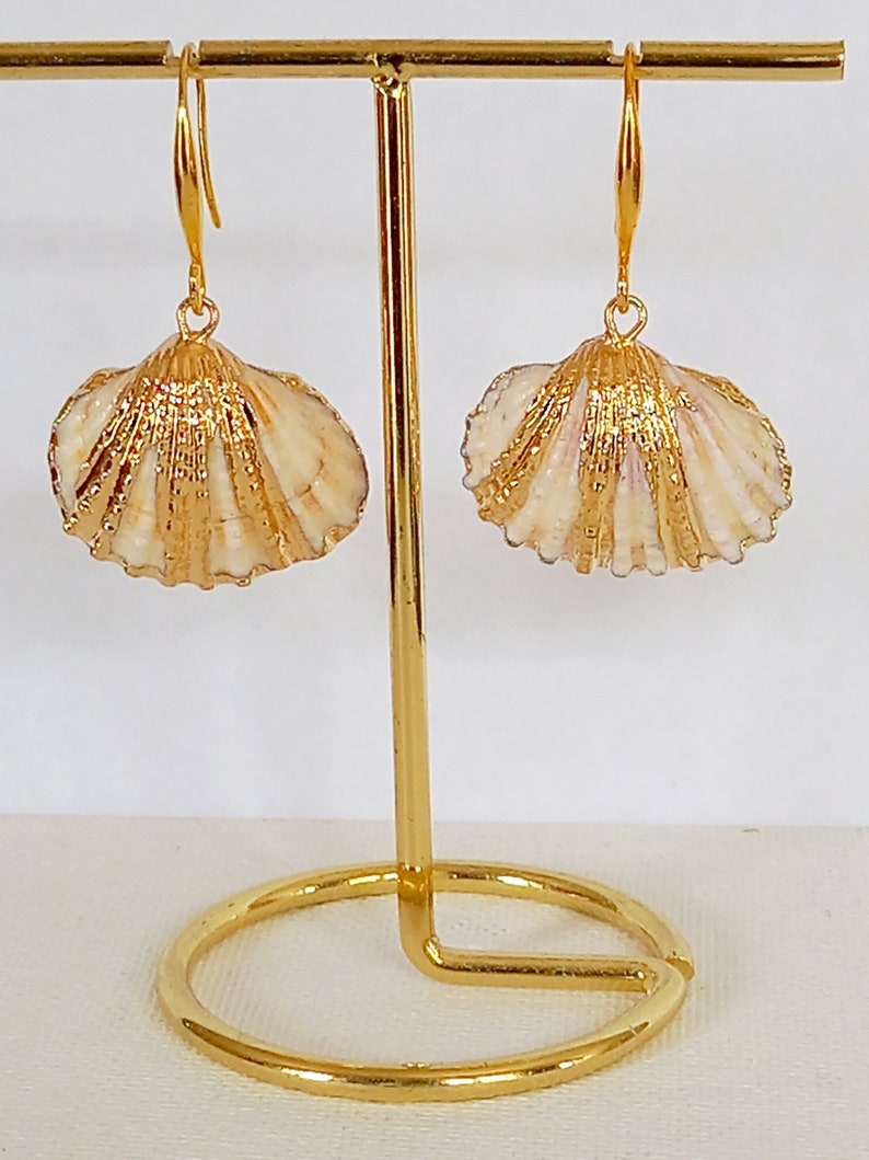 May include: A pair of gold-tone earrings featuring a seashell design. The earrings are hanging from a gold stand.