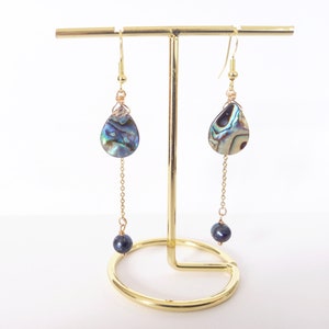 Op de afbeelding: Een paar gouden oorhangers met een traanvormige abalone-schelp en een zwarte parel aan het einde van elke ketting.