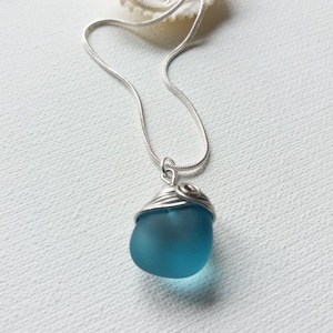 May include: A handcrafted necklace featuring a smooth, teal-colored sea glass pendant wrapped in silver wire. The pendant hangs from a delicate silver chain. The necklace is displayed on a white surface with a seashell in the background.