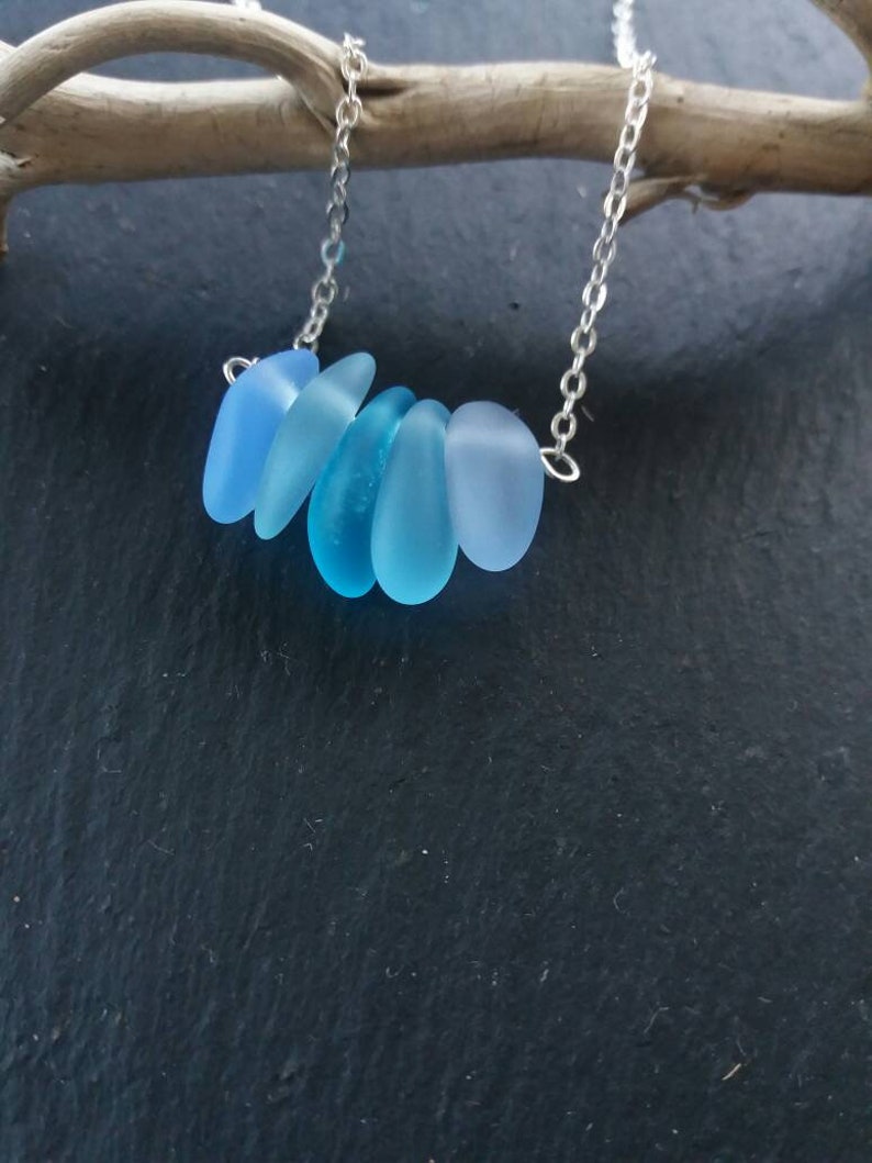 Op de afbeelding: Een zilveren ketting met zes blauwe zeeglas hangers in verschillende tinten blauw. De druppelvormige hangers zijn in een rij gerangschikt. De ketting wordt getoond tegen een donkere achtergrond.