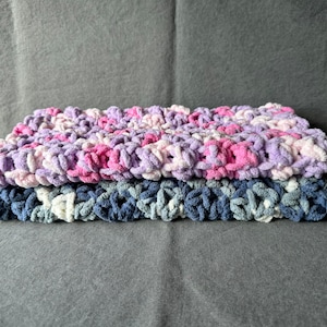 May include: A stack of three crocheted blankets in shades of pink, purple, white, and blue. The top blanket is pink and purple, the middle blanket is white and blue, and the bottom blanket is blue.