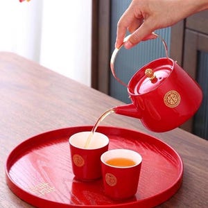 Könnte beinhalten: Rotes Teeservice mit einer Teekanne, die Tee in zwei passende Tassen auf einem roten Tablett gießt. Die Teekanne und die Tassen haben goldene Akzente und ein dekoratives Emblem. Das Teeservice steht auf einem Holztisch.