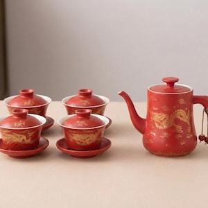 May include: A red ceramic tea set with gold dragon designs. The set includes a teapot with a wooden handle and four teacups with saucers. The teapot has a long spout and a lid. The teacups have lids.