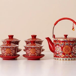 May include: A red and gold tea set featuring a teapot and several lidded teacups with saucers. The teapot has a curved handle and spout. The set is decorated with floral and Chinese character designs.