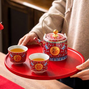 May include: A red ceramic tea set with a teapot and two cups, all adorned with a gold emblem and floral patterns. The set sits on a matching red tray. The teapot has a gold handle and a decorative lid. The cups are filled with tea.