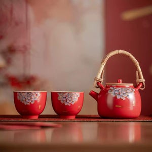 May include: A red ceramic teapot with a woven rattan handle and two matching red ceramic tea cups. Each piece features a painted light purple and white lotus flower design with gold accents. The teapot has a lid and a short, curved spout.