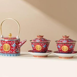 Puede incluir: Un juego de té rojo y dorado con una tetera y dos tazas de té. La tetera tiene un asa y una tapa doradas, y las tazas tienen tapas y platillos a juego. El juego presenta un diseño floral y caracteres chinos.