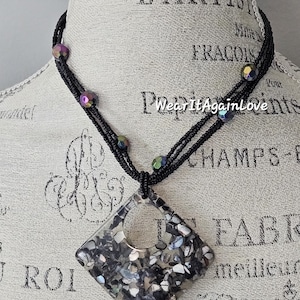 May include: A black beaded necklace with a square pendant made of iridescent shell pieces. The pendant has a circular cutout in the center.