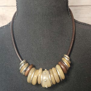 May include: A brown leather necklace with a series of beige, brown, and silver beads. The beads are various sizes and shapes, creating a unique and stylish accessory. The necklace is displayed on a black mannequin.