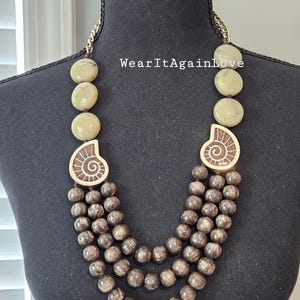 May include: A multi-strand necklace with brown wooden beads and two large beige shell-shaped beads with gold accents. The necklace is on a mannequin.