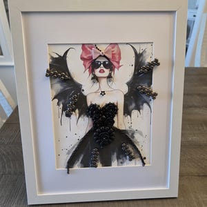 May include: A framed mixed-media artwork featuring a watercolor illustration of a figure with black wings, wearing sunglasses and a pink bow. The figure is adorned with three-dimensional black floral embellishments and metallic studs on the wings.