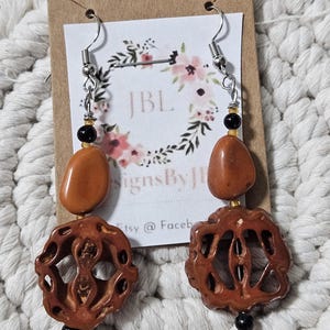 May include: A pair of handmade earrings featuring a combination of brown, orange, and black beads. The earrings have a carved wooden element and a smooth, oval-shaped stone. The earrings are suspended from silver-colored ear wires.