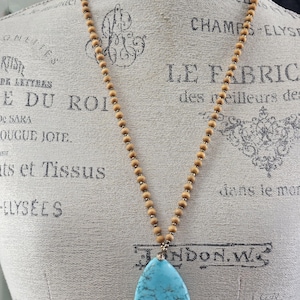 May include: A turquoise teardrop pendant necklace with a wooden bead chain. The necklace is displayed on a mannequin.