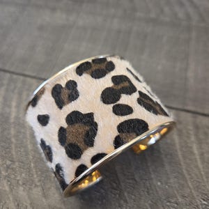 May include: A gold-toned metal cuff bracelet with a leopard print design. The bracelet features a cream-colored background with brown and black spots, and a polished gold finish.