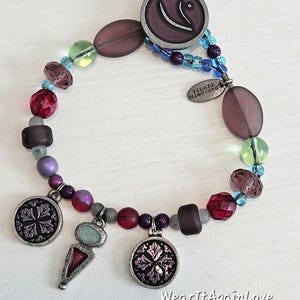 May include: A multi-colored beaded bracelet with silver charms. The charms feature a floral design and a purple and silver swirl. The bracelet has a silver tag that reads "Danforth Jewelry".