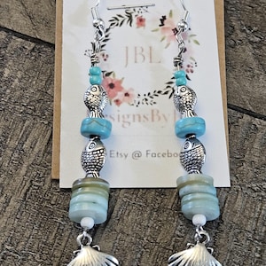 May include: Dangling earrings with a beach theme. Each earring features a silver fish charm, turquoise beads, and a silver seashell pendant. The earrings are suspended from silver hooks and are displayed against a floral design.