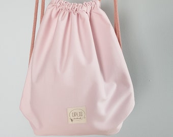 Könnte beinhalten: Ein rosa Rucksack mit Kordelzug und einem weißen Logo mit der Aufschrift "Tipi.eu" und einem kleinen Pfeil. Der Rucksack hängt an einem weißen Schaukelpferd auf einem Regal.