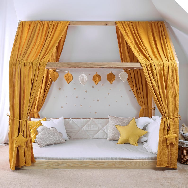 May include: A wooden bed frame with a canopy draped in mustard yellow fabric. Decorative leaf and star-shaped accents adorn the bed. Pillows in various shapes and colors are arranged on the bed.