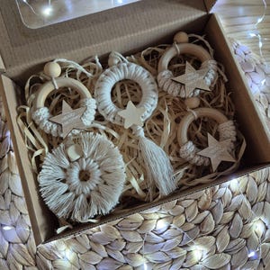 Op de afbeelding: Een verzameling handgemaakte kerstversieringen in een kartonnen doos. De ornamenten zijn wit en beige, met houten ringen, sterren en macramé details. Sommige ornamenten hebben de tekst "MERRY CHRISTMAS". De doos staat in een gevlochten mand.