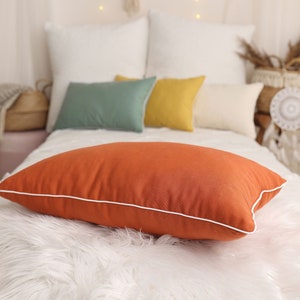 May include: A burnt orange throw pillow with a white border sits on a white faux fur throw blanket. The pillow is on a white bed with other pillows in green, yellow, and white.