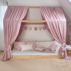 May include: A light wood canopy bed with pink curtains and pillows. The bed frame is rectangular, and the curtains are draped over the top. Decorative leaf-shaped ornaments hang from the frame. Several pink and white pillows are arranged on the bed.