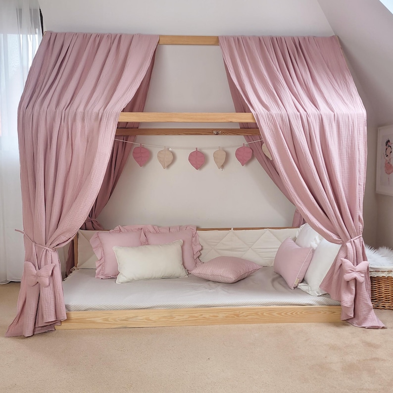 May include: A light wood canopy bed with pink curtains and pillows. The bed frame is rectangular, and the curtains are draped over the top. Decorative leaf-shaped ornaments hang from the frame. Several pink and white pillows are arranged on the bed.