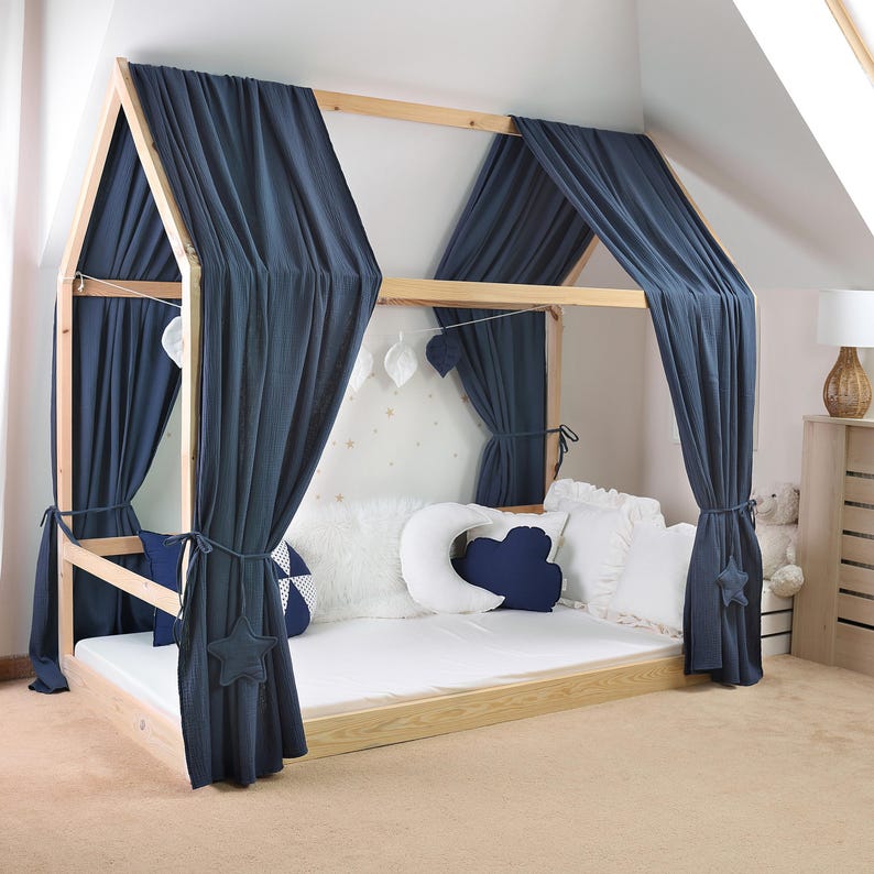May include: A wooden house-shaped bed frame with navy blue curtains. The bed is decorated with white and navy blue pillows and star-shaped accents. The bed frame is made of light-colored wood.