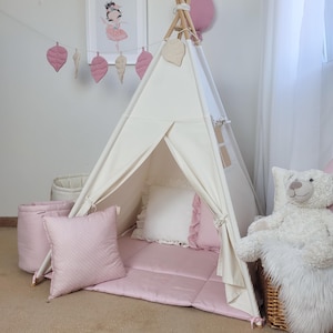 Può includere: Una tenda teepee color crema con pali di legno, posizionata su un tappetino trapuntato rosa. La tenda è decorata con cuscini rosa, un orsacchiotto bianco e una ghirlanda rosa con forme di foglie. Una stampa di ballerina incorniciata è appesa al muro.