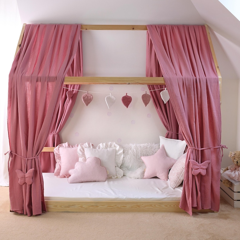 May include: A wooden framed bed with pink curtains and decorative pillows. The bed frame has a canopy design with hanging leaf-shaped ornaments. The bed is decorated with pink and white pillows, including a star-shaped pillow and a cloud-shaped pillow.