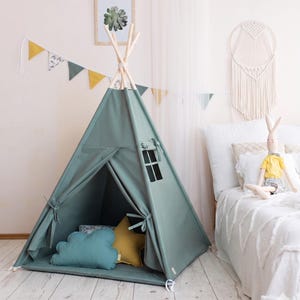 Könnte beinhalten: Ein salbeigrünes Stoff-Tipi mit einem Holzstangengestell, einem Seitenfenster mit Bindevorhängen und einem Vordereingang, der offen gebunden werden kann. Im Inneren befinden sich ein blaues wolkenförmiges Kissen und ein senfgelbes sternförmiges Kissen.