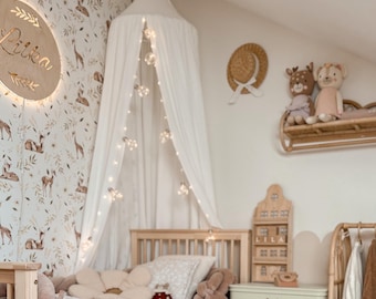 Könnte beinhalten: Ein helles Kinderzimmer mit einem Holzbett, einem weißen Baldachin mit Lichterketten und einer Tapete mit floralem Muster. Das Zimmer ist mit Stofftieren, einem Holzschild mit der Aufschrift "Lilia" und einem kleinen Holzregal dekoriert.