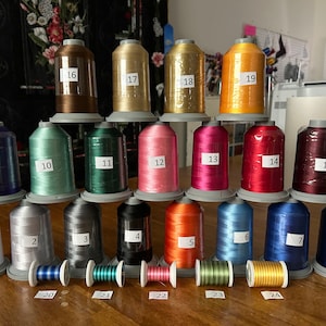 Op de afbeelding: Een verzameling van 24 spoelen garen in verschillende kleuren, genummerd van 1 tot en met 24. De spoelen zijn in een piramidevorm op een houten oppervlak gerangschikt.