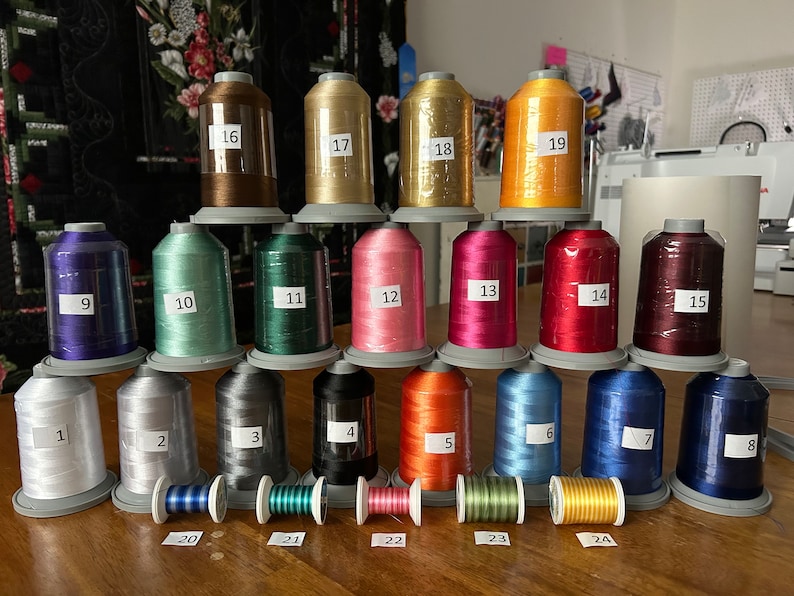 Op de afbeelding: Een verzameling van 24 spoelen garen in verschillende kleuren, genummerd van 1 tot en met 24. De spoelen zijn in een piramidevorm op een houten oppervlak gerangschikt.