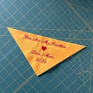 Op de afbeelding: Een gele driehoekige stof met rode geborduurde tekst die "You are my sunshine, Love, Mom 2024" en een rood hart zegt.
